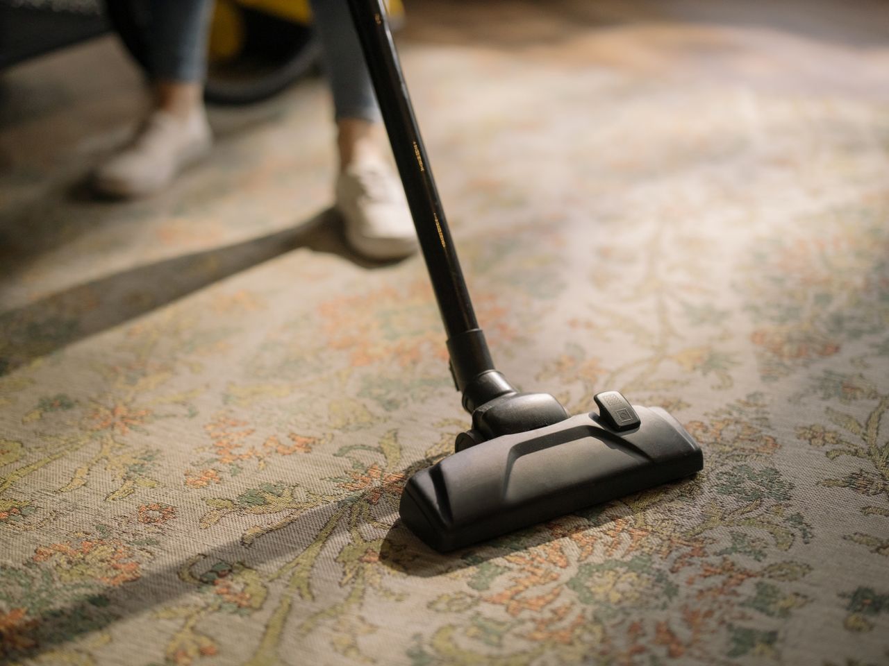 Gros plan d’un aspirateur posé sur un tapis à motifs dans une pièce baignée de soleil, illustrant un instant de nettoyage domestique.